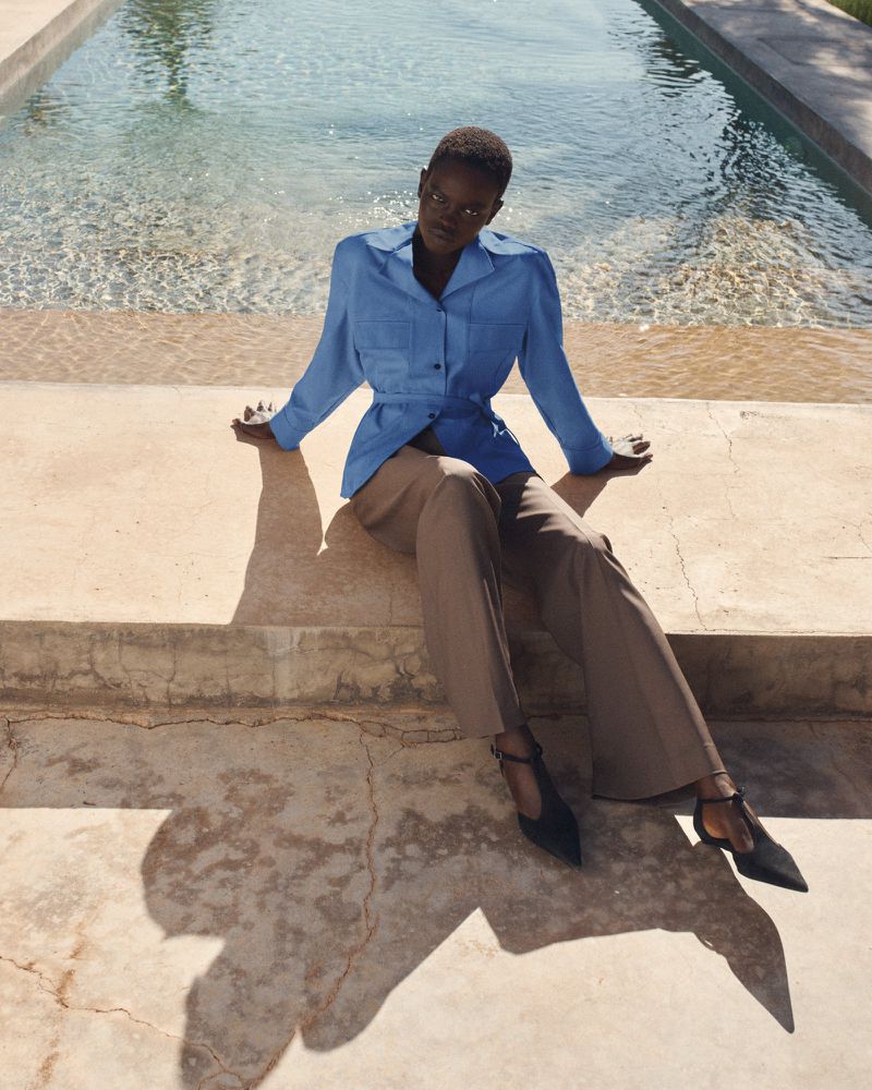 Adult woman in blue jacket and tan trousers sits on steps near pool in an outdoor setting with natural lighting and shadows.