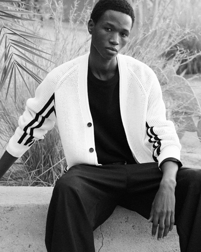 Black and white portrait of a young adult man in a knitted cardigan and dark pants, sitting outdoors with palms in the background.