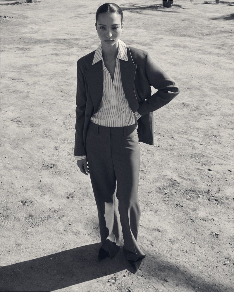 Model wearing a structured blazer, striped shirt, and trousers outdoors on a sandy ground for COS lookbook.