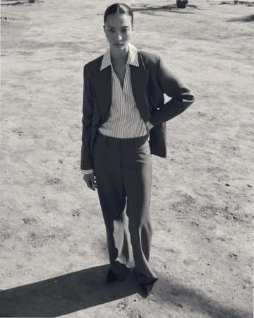 Model wearing a structured blazer, striped shirt, and trousers outdoors on a sandy ground for COS lookbook.