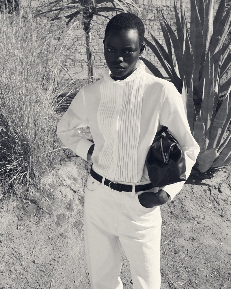 Black-and-white fashion portrait of a model wearing a ruffled white shirt and holding a dark handbag outdoors.