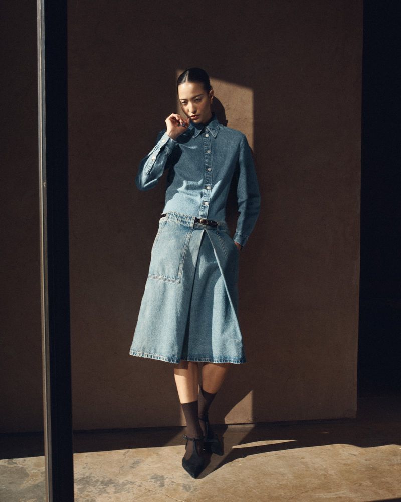 Model wearing a blue denim shirt and matching skirt standing in sunlight against a dark wall for COS lookbook