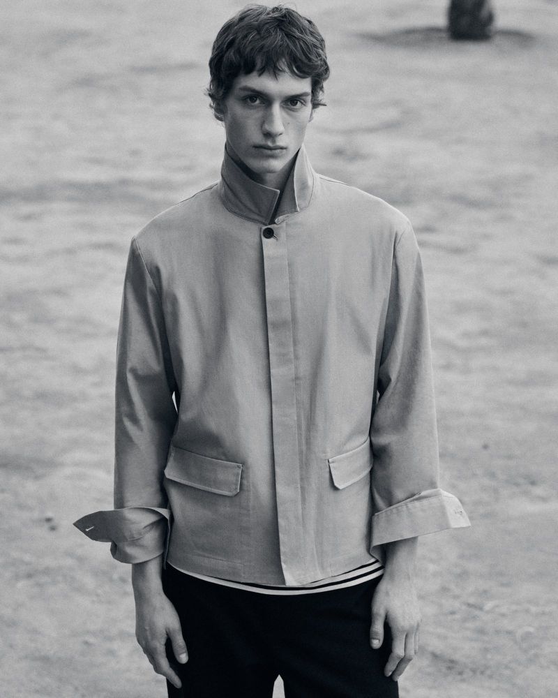 Monochrome portrait of a young adult man in a jacket, standing outdoors in natural light. Fashion editorial style, neutral tones.