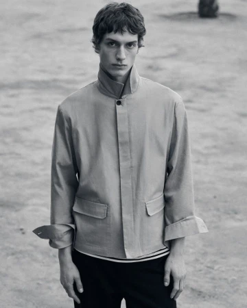 Monochrome portrait of a young adult man in a jacket, standing outdoors in natural light. Fashion editorial style, neutral tones.