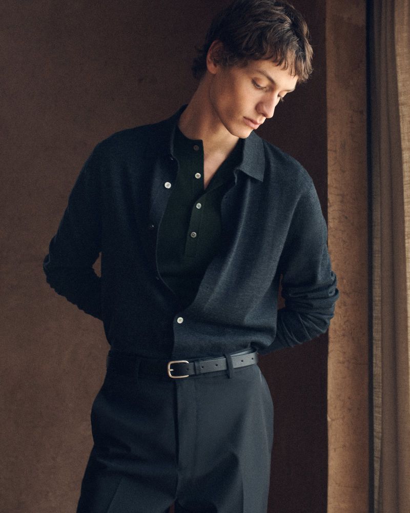A fashionable young man in a navy collared sweater poses indoors against a brown wall, with a natural light accentuating his features.