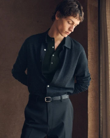 A fashionable young man in a navy collared sweater poses indoors against a brown wall, with a natural light accentuating his features.