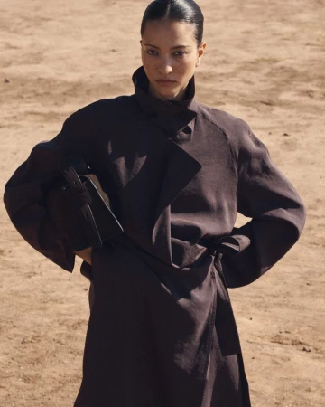 Model in a dark brown coat with a high collar holding a matching handbag against an earthy outdoor backdrop.