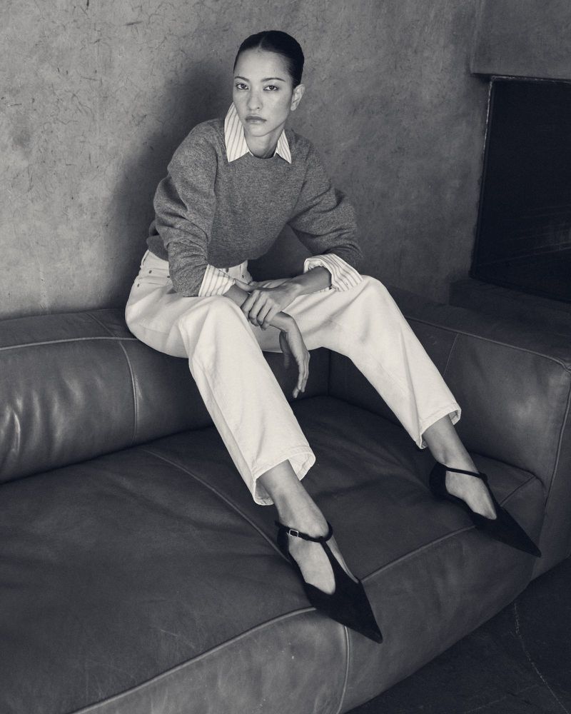Model seated on a leather sofa wearing a grey sweater, striped shirt, light trousers, and black T-strap shoes.