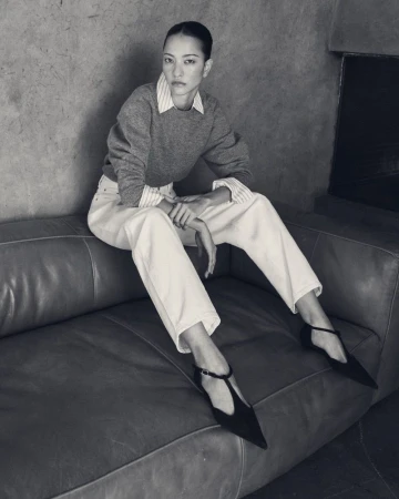 Model seated on a leather sofa wearing a grey sweater, striped shirt, light trousers, and black T-strap shoes.