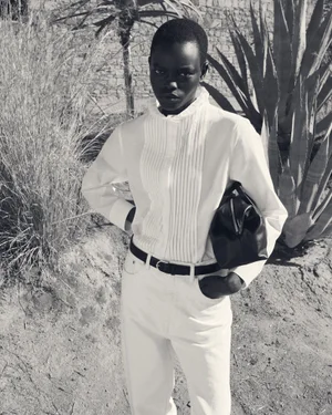Black-and-white fashion portrait of a model wearing a ruffled white shirt and holding a dark handbag outdoors.