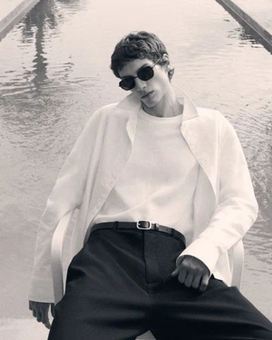 Man wearing sunglasses and white shirt seated near an outdoor pool for COS Pre-Spring 2026