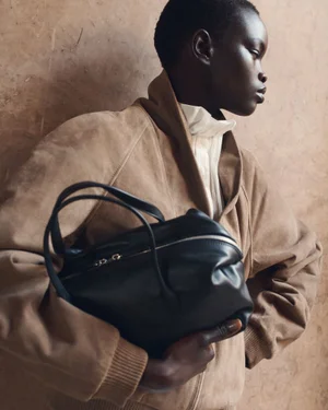 Model with short hair wearing a beige suede bomber jacket holding a black leather handbag against a textured brown wall.