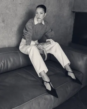 Model seated on a leather sofa wearing a grey sweater, striped shirt, light trousers, and black T-strap shoes.