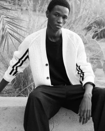 Black and white portrait of a young Black man seated outdoors wearing a white cardigan and dark trousers for COS Pre-Spring 2026 lookbook.