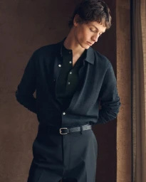 Model wearing dark buttoned polo shirt and trousers with belt against a textured wall for COS lookbook.