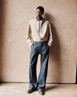 Man wearing a beige button-down shirt and blue denim jeans posing against a textured wall