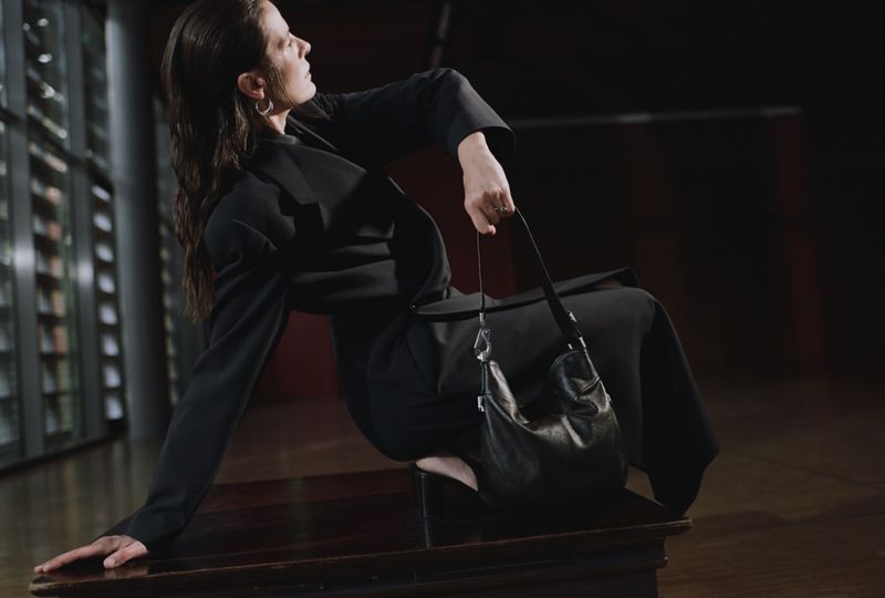 Woman in black suit sits on a wooden surface holding a black leather purse in a dimly lit indoor setting with modern architecture.