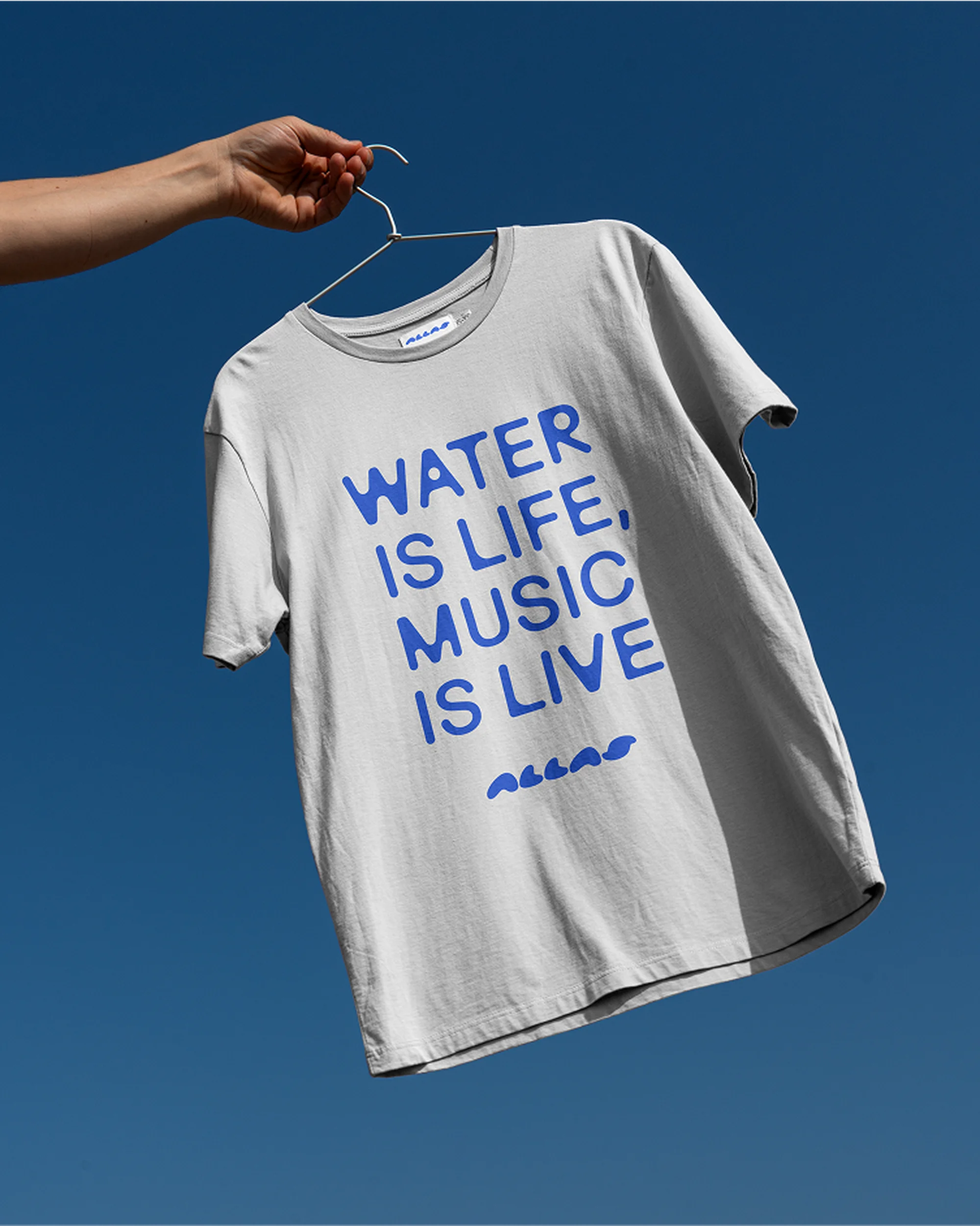 Grey t-shirt on a hanger with blue text 'Water is Life, Music is Live' against a clear blue sky. Fashion apparel shot.
