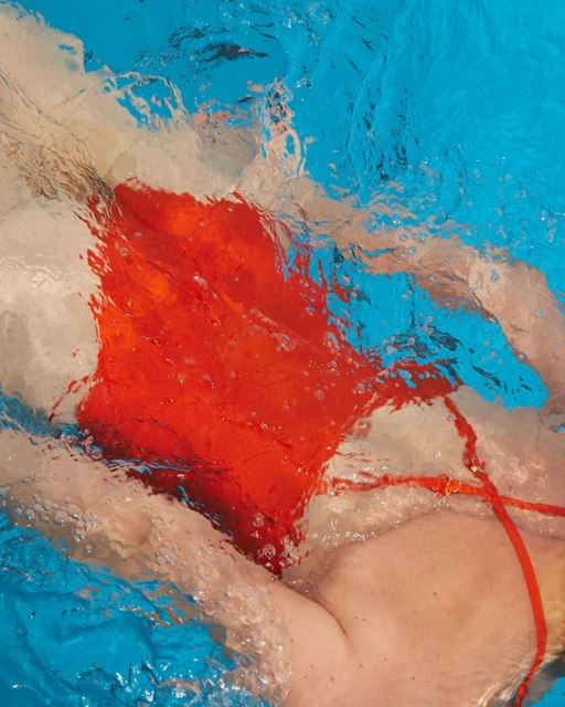 Underwater view of a person in a red swimsuit swimming in a pool with rippling water and blue pool tiles visible through the water.