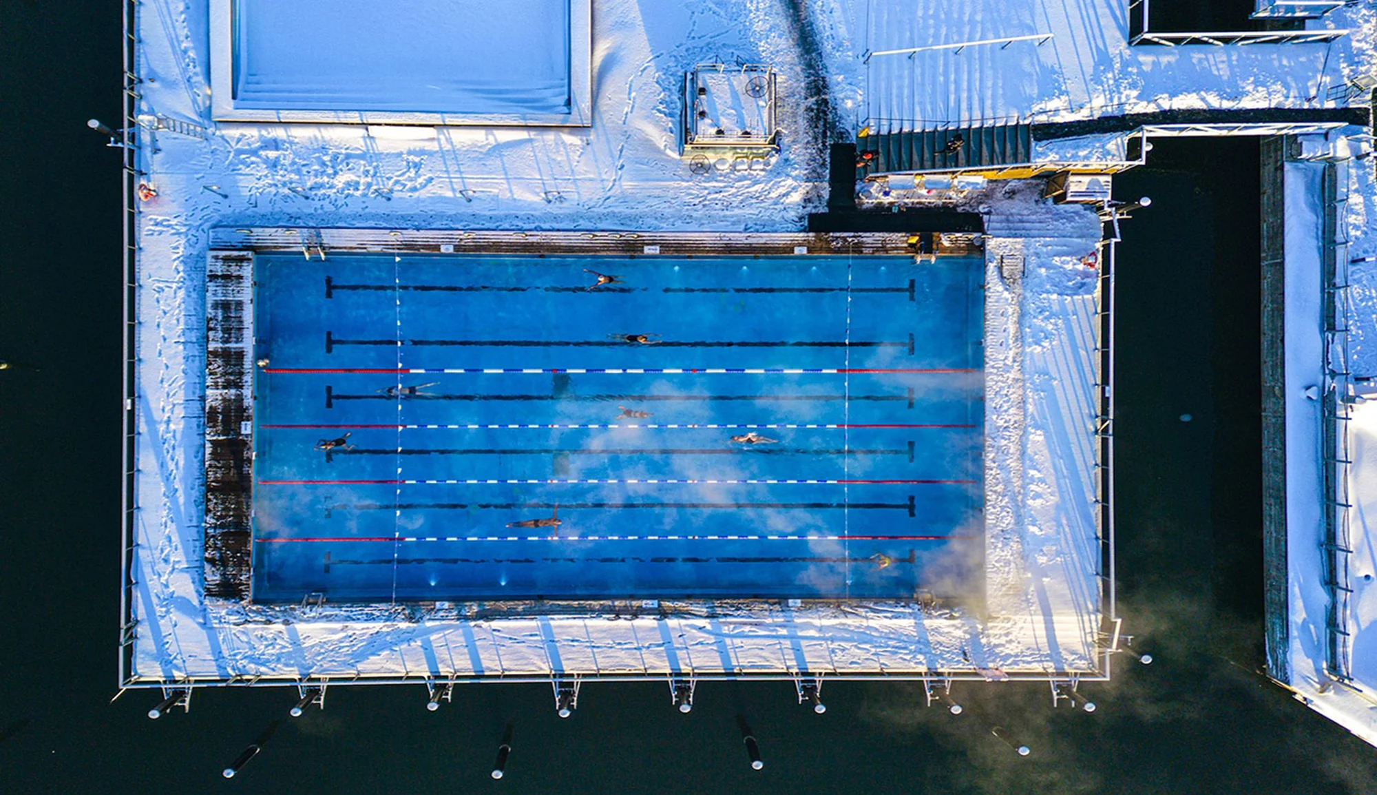Aerial view of swimmers in an outdoor pool with snow-covered edges and surrounding architecture, highlighting winter sports and recreation.