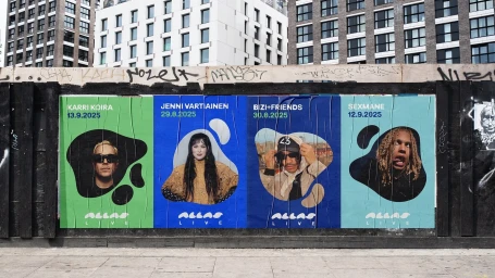 Four colorful concert posters pasted on a textured outdoor wall beneath modern apartment buildings.