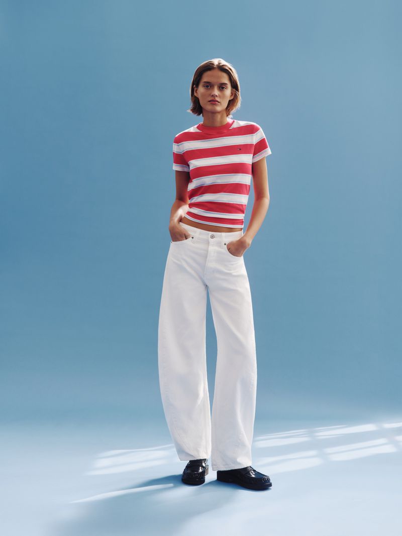 Full-length shot of a woman posing in a red and white striped tee shirt, white pants, and black shoes against a solid blue background.
