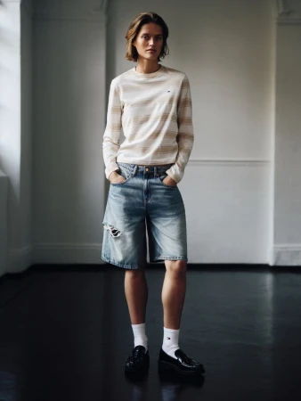 Adult female in a photo studio wearing a long-sleeve shirt, jean shorts, white socks, and black loafers poses with hands in pockets.