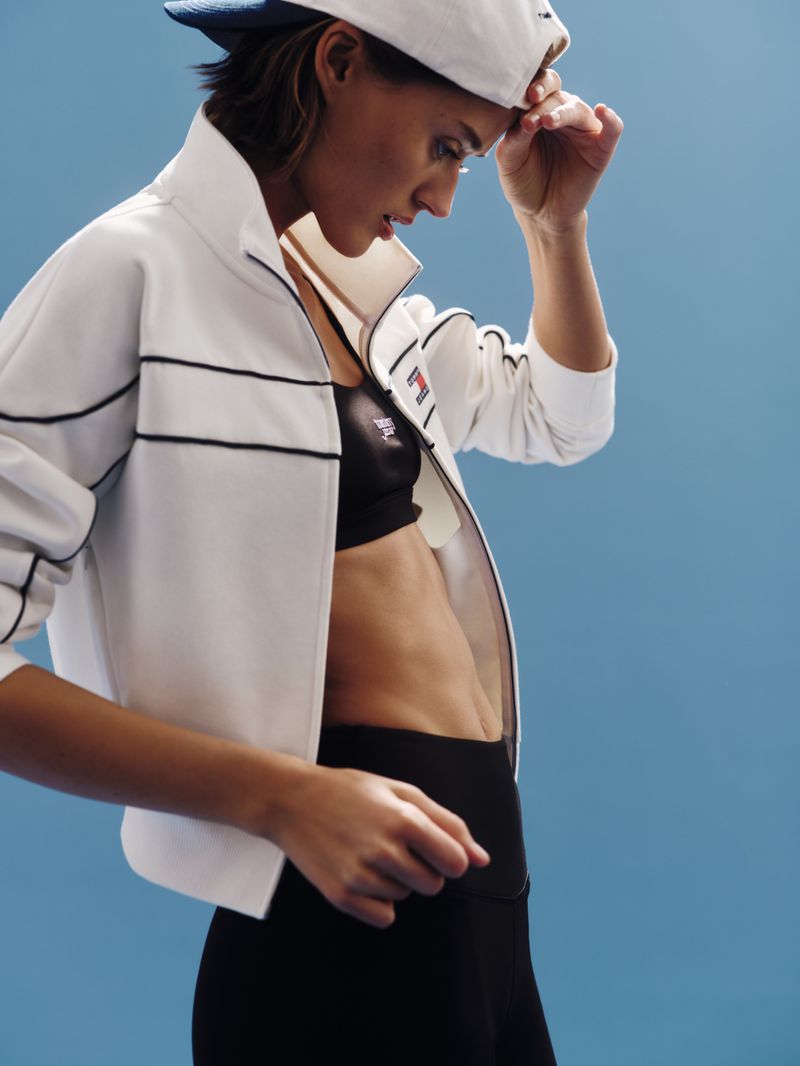 Attractive woman in sportswear: white zip-up jacket, black sports bra, and leggings, wearing a cap and posing against a blue backdrop.