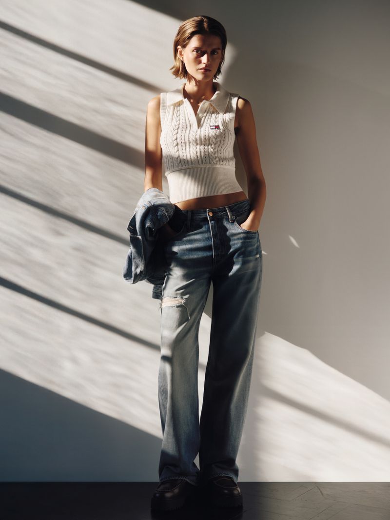 A fashionable woman posing in a sleeveless knit top and denim jeans stands against a sunlit white wall, creating a striking visual.