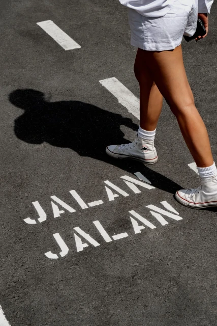 Woman walks on asphalt road with white 'Jalan Jalan' text, wearing white sneakers, shorts, and top in outdoor setting with shadow.