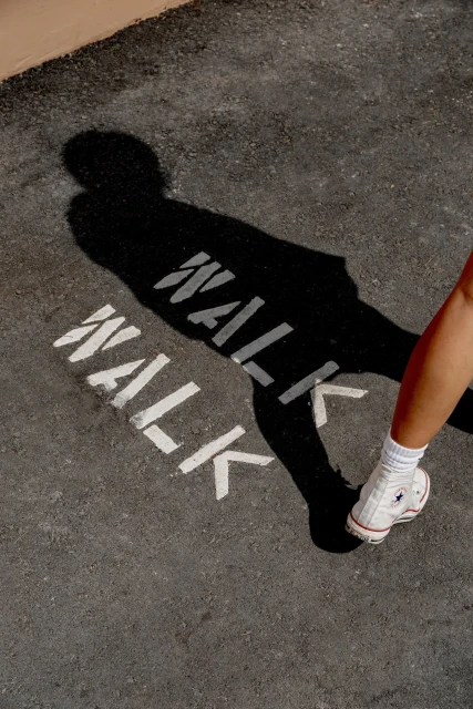 Low angle shot of a person wearing white sneakers standing on asphalt next to 'WALK' marking, with the figure's shadow present.