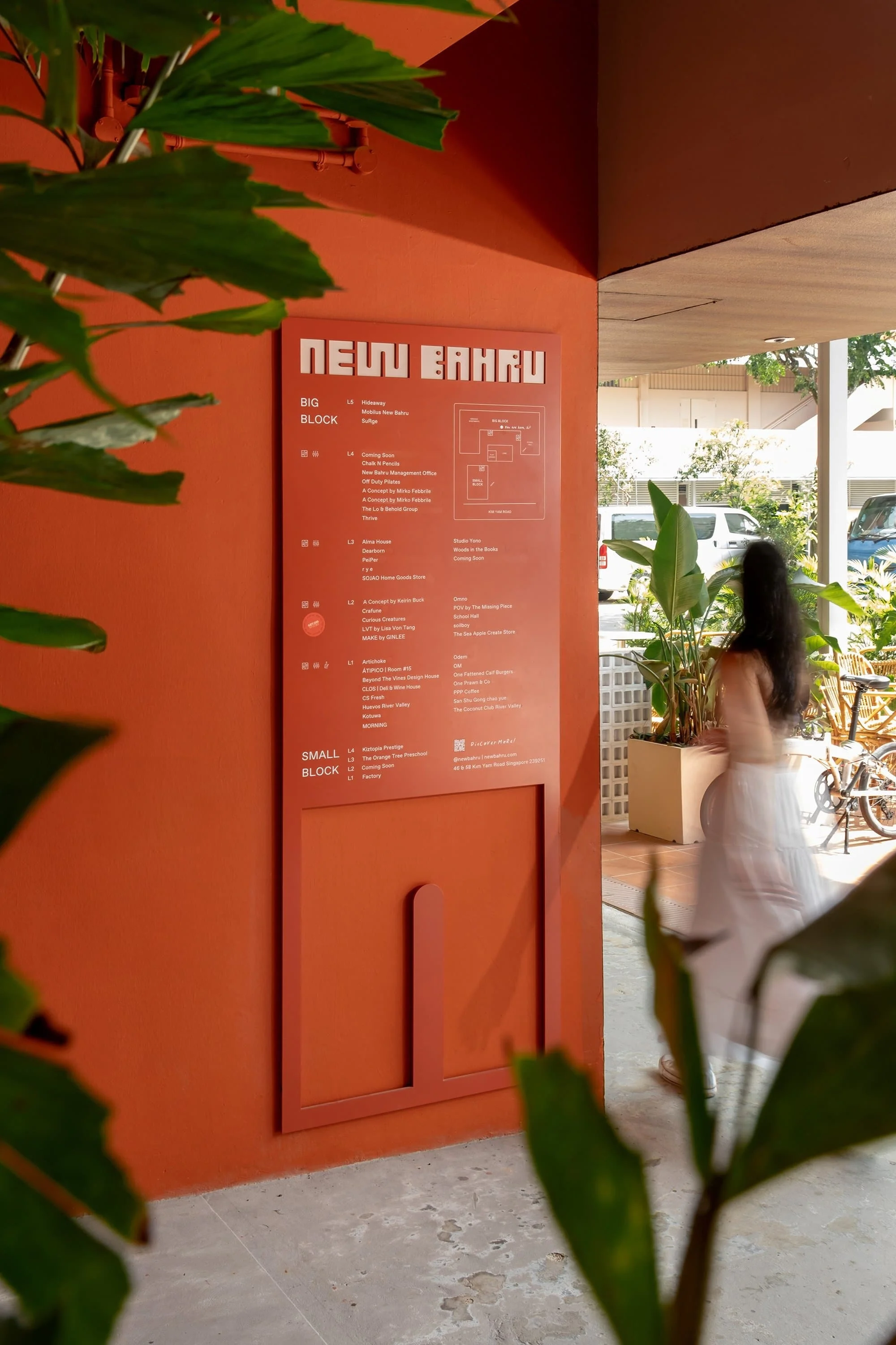 Architectural design featuring an orange wall with a directory sign, a blurred woman in a white dress, and green plants in an urban setting.