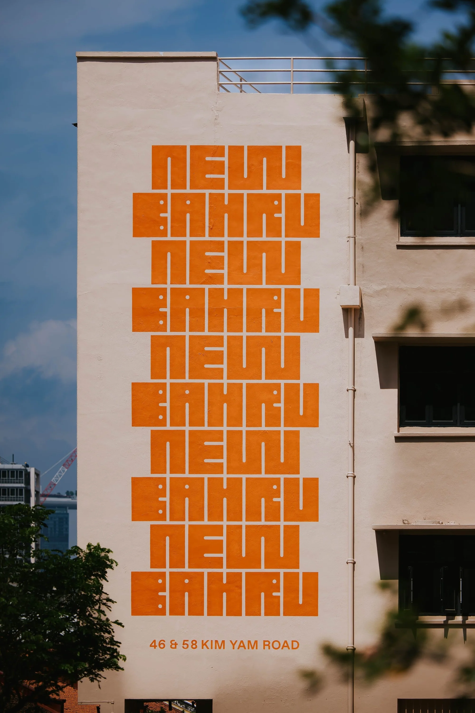 Exterior view of a building with large, orange graphic text spelling 'NEAU BAHRU' and an address on a white facade under a blue sky.
