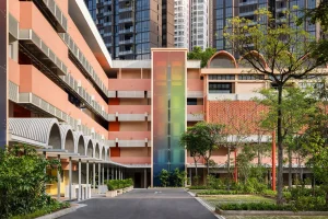 Modern pink multi-story building exterior with a central iridescent glass elevator tower and arched structural awnings.