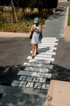 Woman in light dress and blue cap walking on a paved pathway with white painted rectangular markings