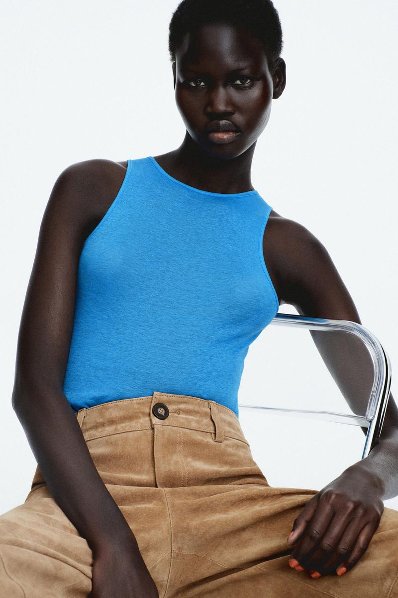 Dark-skinned woman in a turquoise tank top poses in front of a white background, sitting on a transparent chair with brown pants.