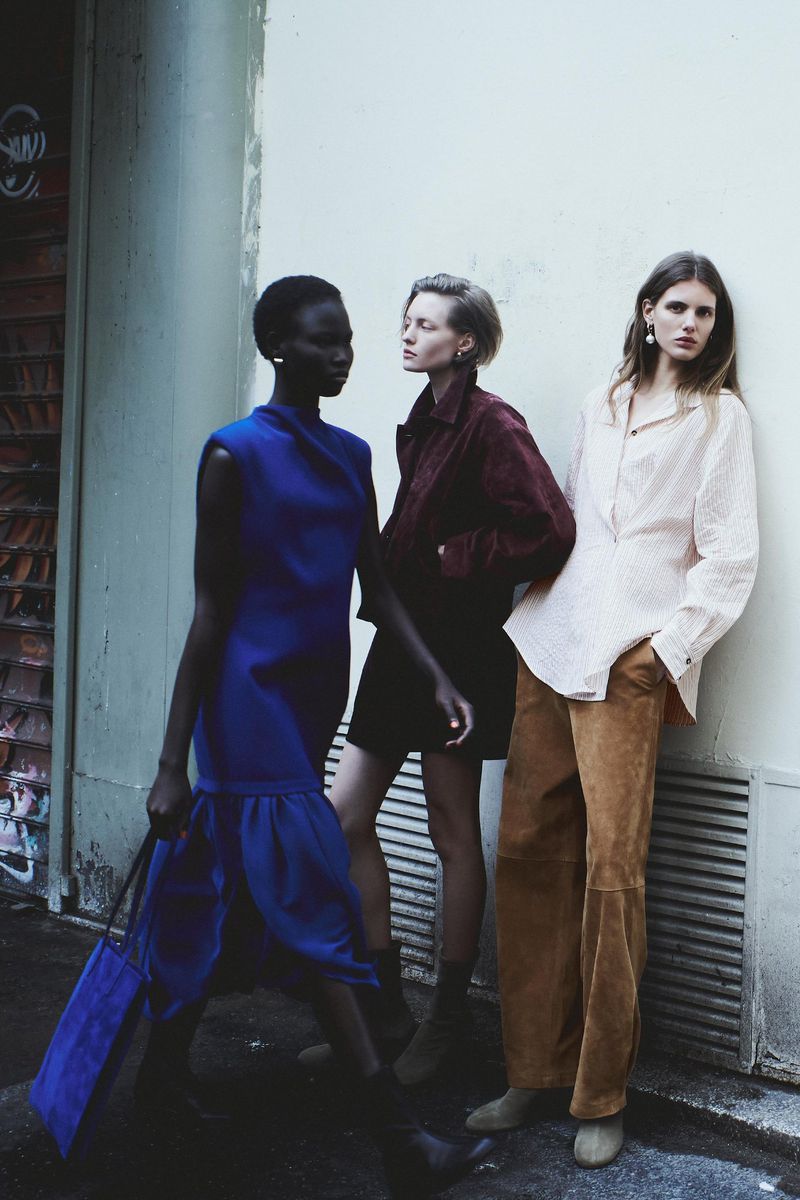 Three fashion models pose against an urban exterior wall wearing a blue dress, maroon jacket and dress, and beige top and pants.