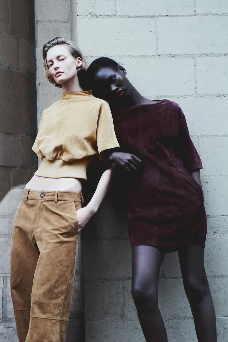 Two women in fashionable outfits pose against a concrete block wall, one in tan separates, the other in a burgundy dress and black stockings.