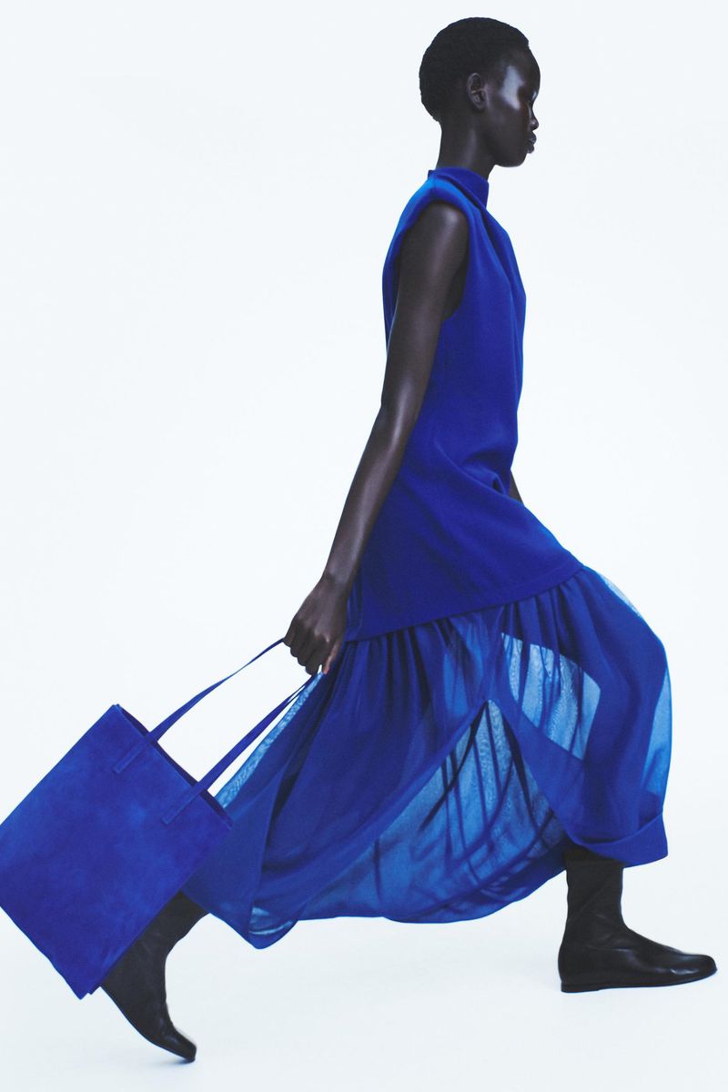 Runway fashion shot: a Black female model walks in profile wearing a cobalt dress, boots, and carrying a matching blue tote bag.