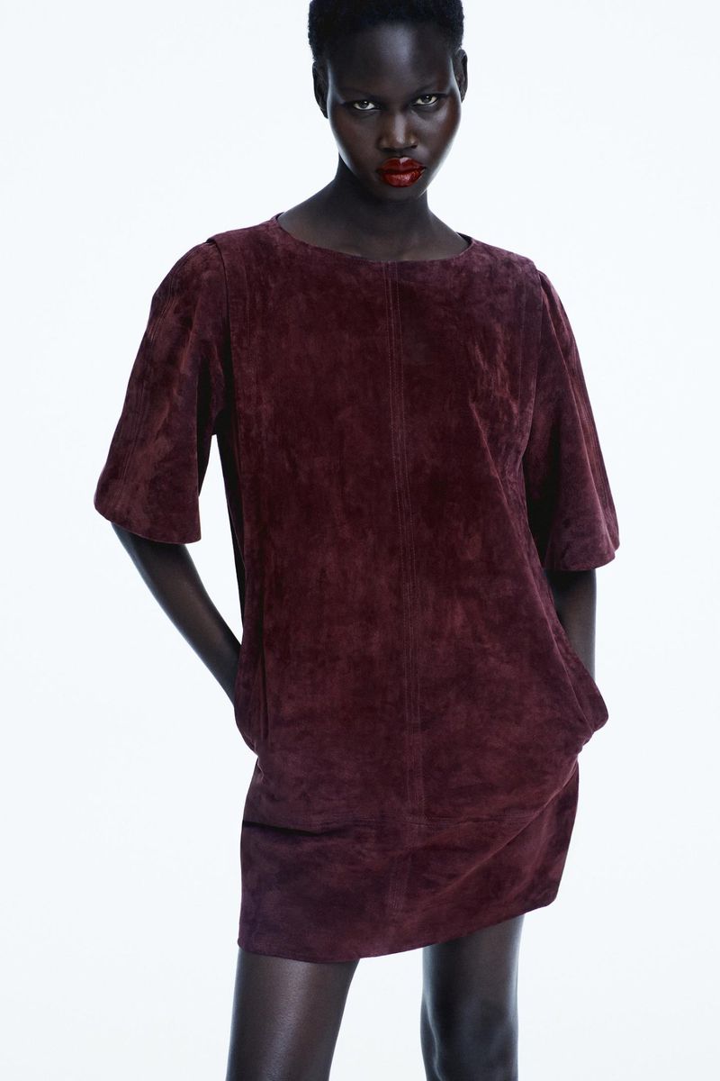 A dark-skinned woman poses in a studio wearing a short-sleeved suede dress. She is facing forward and is slightly angled to the right.