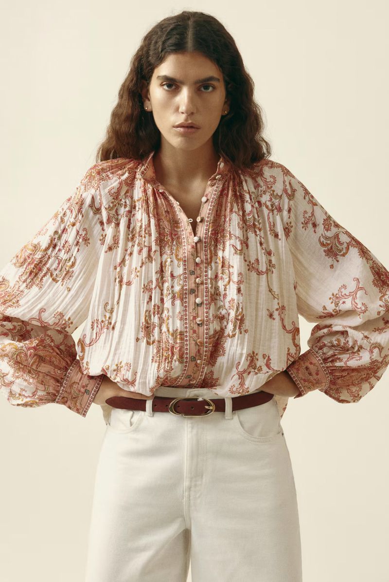 Fashion shot of a woman wearing a white and coral print blouse with balloon sleeves and white jeans against a beige background.