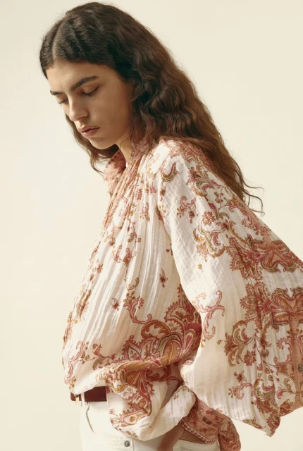 A woman with long wavy brown hair poses in a white paisley print blouse with a brown belt and white pants against a cream background.
