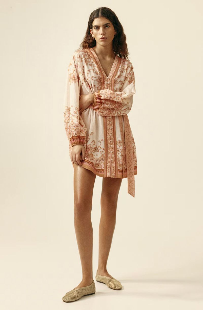 A full-length studio shot of an adult woman in a patterned dress, accented by long sleeves and a waist tie, posing against a neutral backdrop.