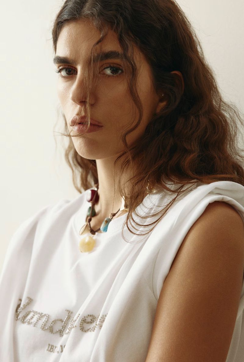 Close-up portrait of an adult woman with curly hair wearing a white sleeveless top and a colorful beaded necklace against a neutral backdrop.