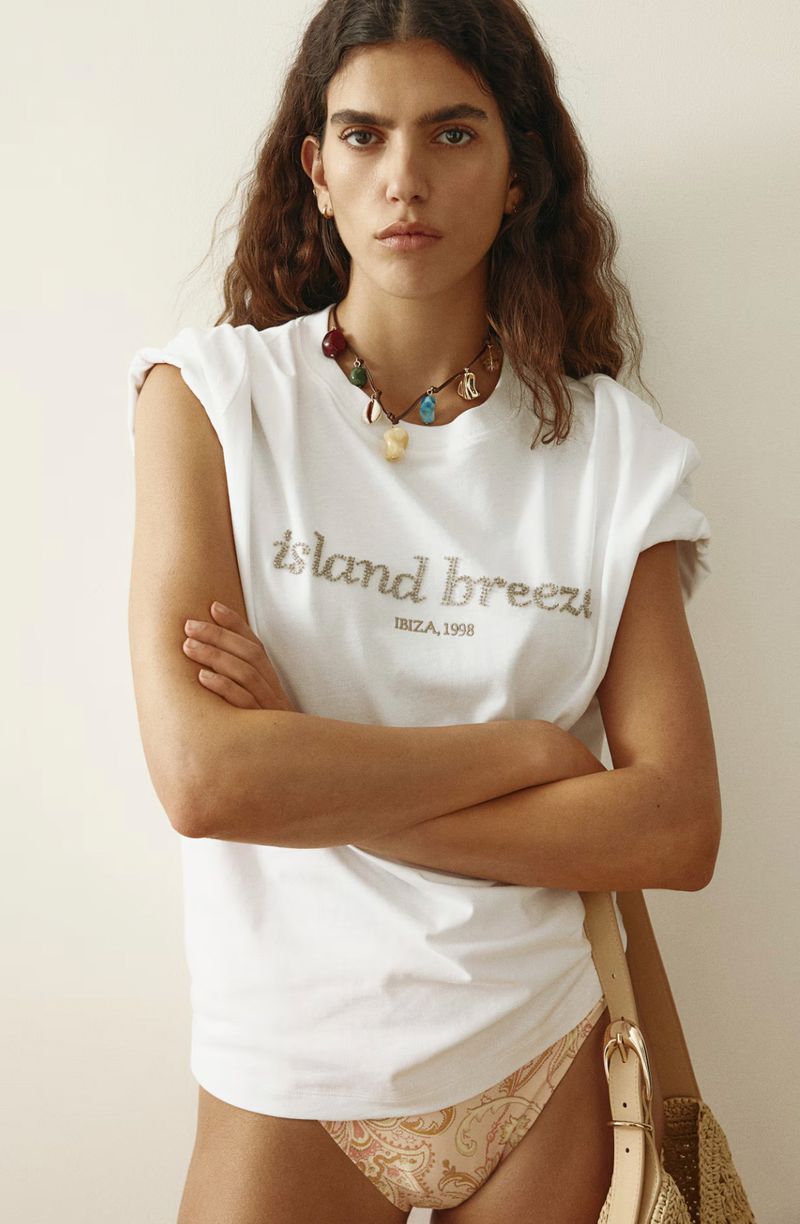 Model wearing a white graphic t-shirt and patterned bikini bottoms with a charm necklace against a plain neutral background.