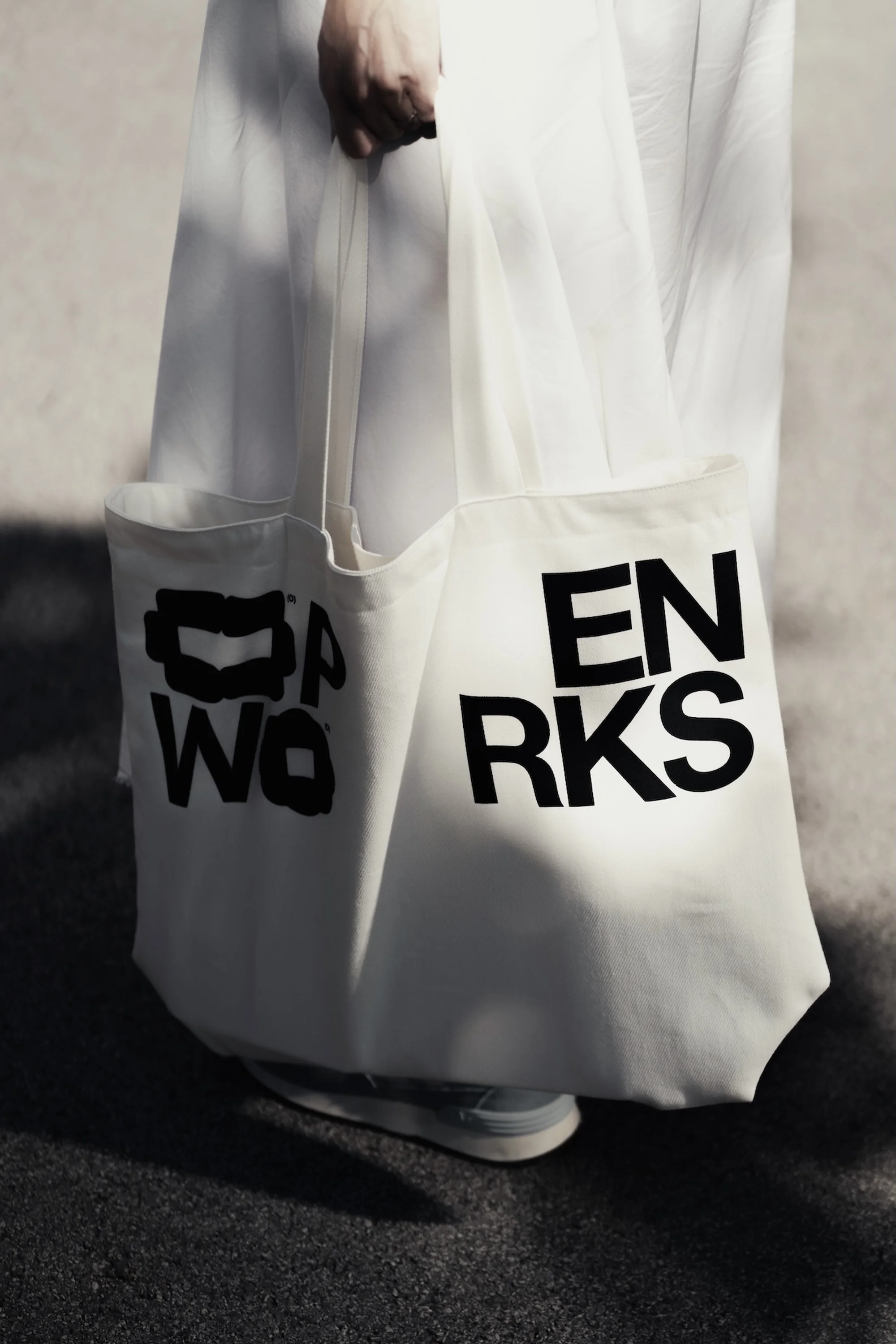 Person holds a white canvas tote bag with black text on a city street, wearing white dress and light-colored sneakers in daylight.