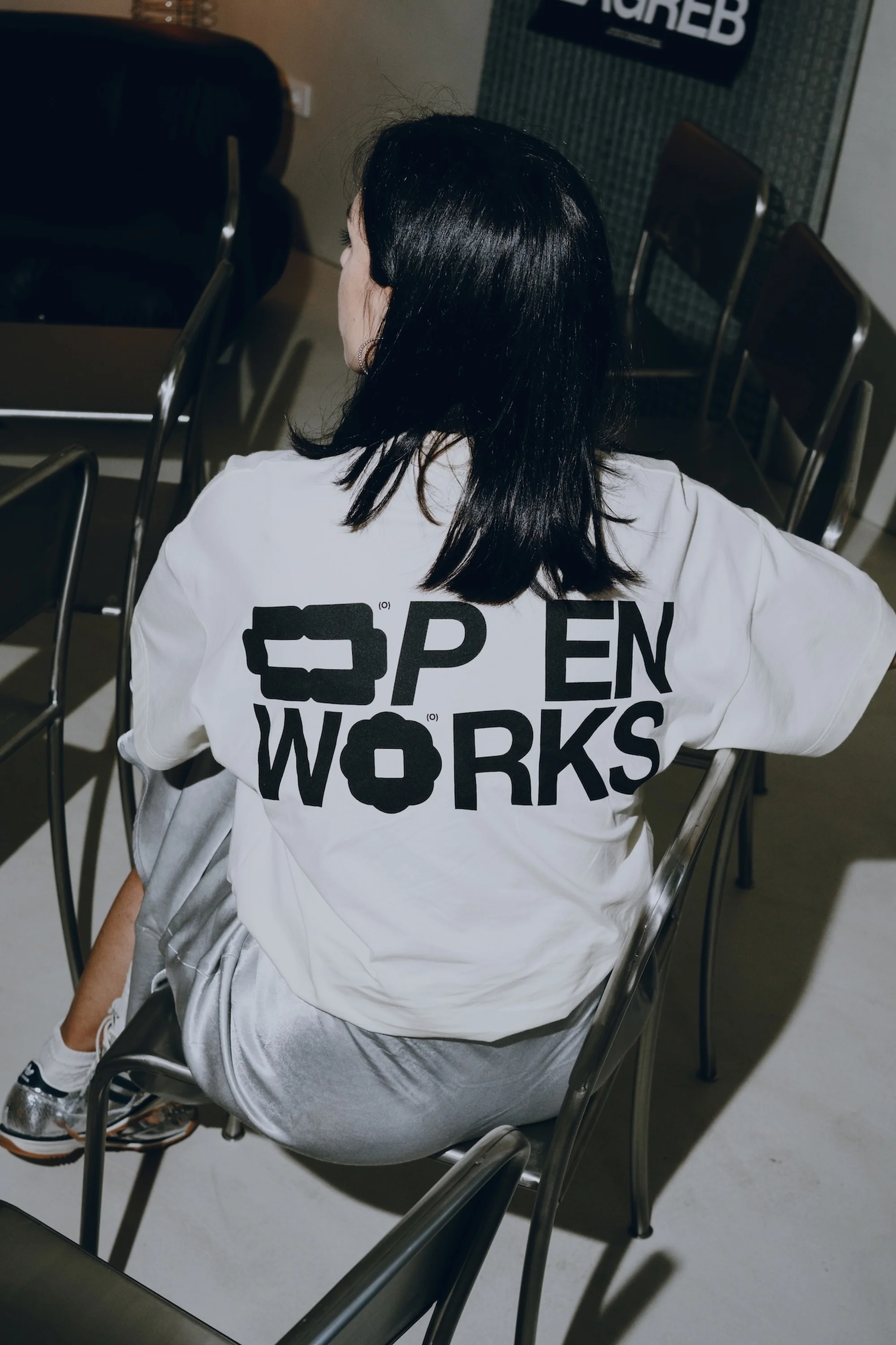 Back view of a woman with black hair wearing a white Open Works t-shirt seated among chairs in a brightly lit indoor setting.