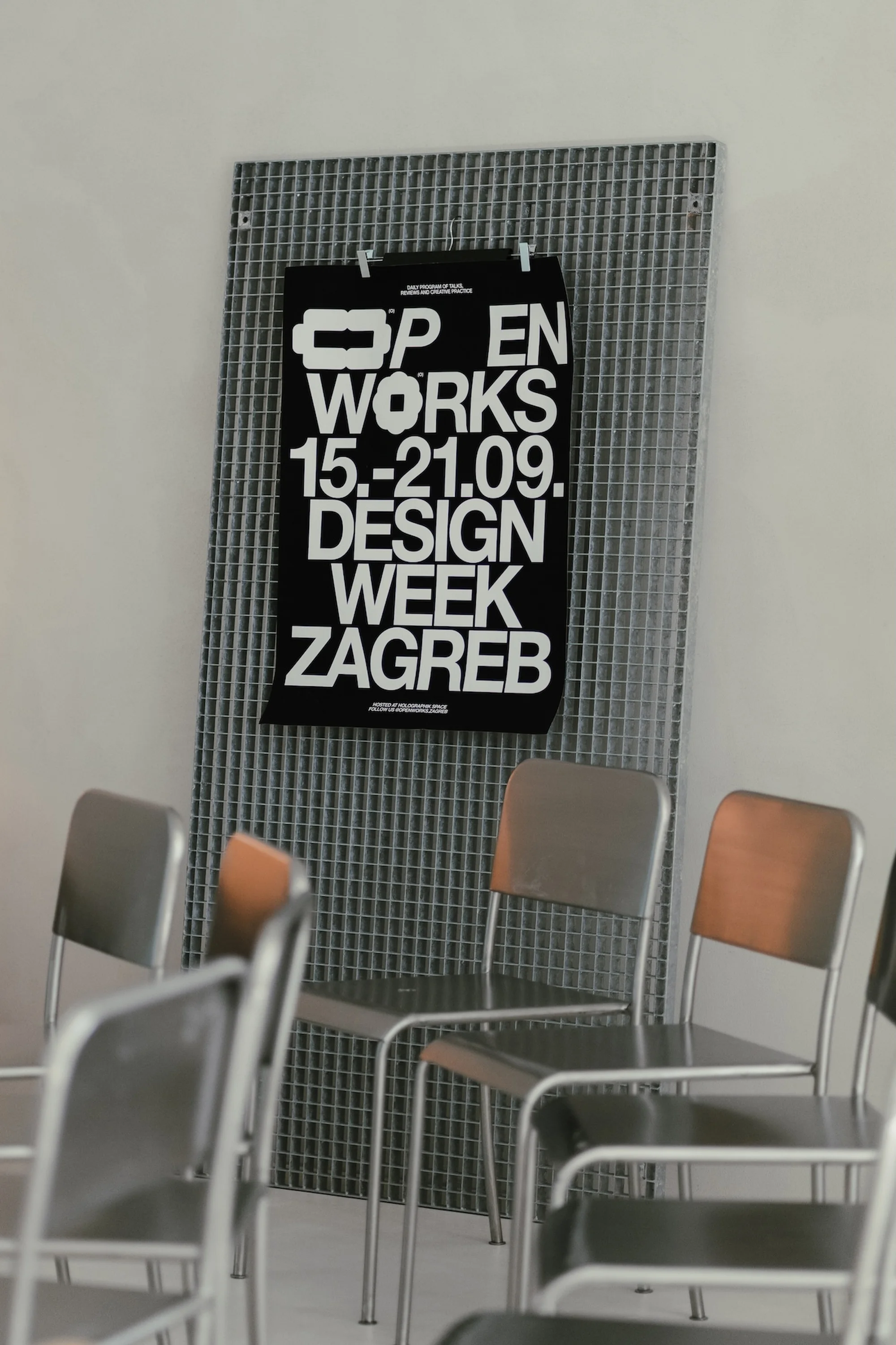 Poster for Design Week Zagreb hangs behind silver metal chairs in an indoor setting with a light gray wall and modern architecture.