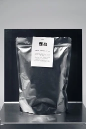 Silver foil pouch with a white label attached to the front set against a grey and black backdrop