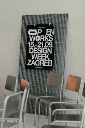 Black poster for Zagreb Design Week on a metallic grid wall behind empty silver metal chairs in a bright interior space.